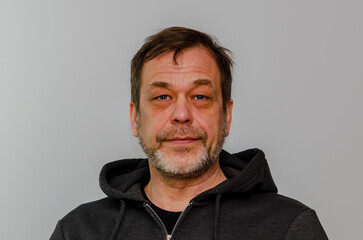 Wall Mural - An elderly 50-55 year old man with a beard wearing a black hoodie looks into the camera against a light colored background.