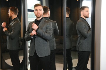 Wall Mural - Modern young handsome businessman dressed in classical suit adjusting a tie in front of the mirror while standing in the suit shop