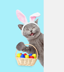 Poster - Happy kitten wearing easter rabbits ears holds basket with colorful easter eggs and looks from behind empty white banner. Isolated on blue background