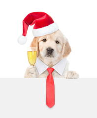 Wall Mural - Young Golden retriever puppy wearing necktie and santa hat looks above empty white banner and holds glass of champagne. isolated on white background