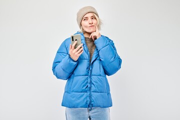 Wall Mural - Portrait of young blond woman in blue jacket looks on mobile phone and thinks. Person with smartphone isolated on white background.
