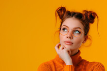 Thoughtful Woman In Orange Turtleneck And Glasses. Pensive Young Woman With Space Buns Wearing Round Glasses And An Orange Sweater, Contemplating Against A Matching Background.