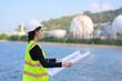 © eakarat - A female chemical engineer stands in front of a chemical tank in a special uniform. Female engineer uses laptop to work outdoors