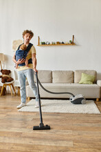 curly father with infant baby boy in carrier vacuuming hardwood floor living room, housework