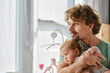 © LIGHTFIELD STUDIOS - curly-haired and pleased father holding in arms his infant boy in cozy bedroom at home, fatherhood