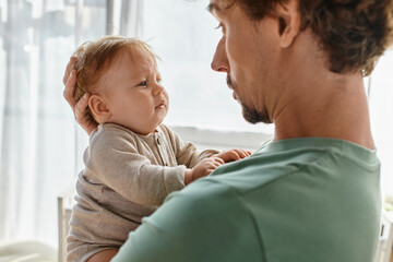 caring father with curly hair and beard holding in arms his infant boy in baby clothes, portrait