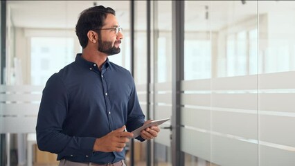 Wall Mural - Smiling professional Indian businessman using tab computer working standing in office. Busy professional business man entrepreneur holding digital tablet tech device working, looking at pad.