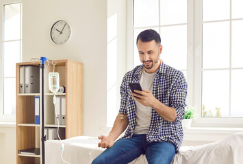 Young man sitting on the couch in clinic using his mobile phone while receiving IV drip infusion and vitamin therapy. Attractive male person in casual clothes receiving injection therapy.