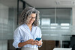 © insta_photos - Smiling mature professional business woman entrepreneur using smartphone in office. Happy older senior female executive, middle aged businesswoman looking at mobile cell phone standing at work.