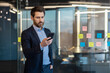© Liubomir - Successful and confident male businessman standing inside office in suit, holding hand in pocket and using mobile phone, reading and sending message, calling, waiting for meeting.