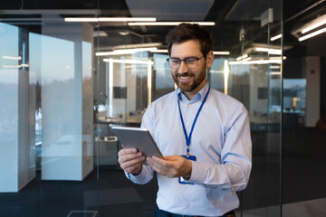 Smiling young man, worker and specialist is standing in the office near the window, holding and using a tablet. Talks online via video call, works with data and projects.