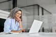 Happy older mature business woman entrepreneur in office using laptop at work, smiling professional senior middle aged female executive wearing glasses working on computer at office workplace desk.