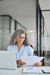 © insta_photos - Busy middle aged business woman working in office with laptop reading document. Mature professional hr manager, notary holding paper finance report sitting at desk in office. Consultancy. Vertical.