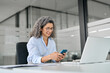 © insta_photos - Smiling mature professional businesswoman executive using smartphone cell mobile apps on cellphone working sitting at desk. Busy middle aged business woman holding phone using cellphone in office.