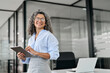 Smiling mature professional business woman bank manager, older happy female executive or lady entrepreneur holding digital tablet pad standing in office at work, looking away at copy space.