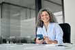 © insta_photos - Happy middle aged senior curly business woman holding phone in office looking at camera. Smiling mature female entrepreneur, older professional worker using cellphone working at desk, portrait.