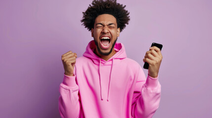 Sticker - excited young man with curly hair in a pink hoodie, holding a smartphone with one hand and making a fist with the other