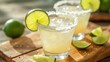  two margaritas sitting on a cutting board with limes and salt on the rim of the glasses and the limes on the cutting board.