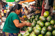 woman buying avocados from a street vendor in a busy market. The vendor is wearing a green shirt, and there are other vendors selling goods nearby