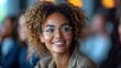 © TheGoldTiger - A smiling woman with curly hair and glasses, in a room with out-of-focus people behind., generative ai