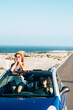 © simona - Travel friends and transport with blue convertible car and couple of adult women friends have fun together driving on a long road with ocean in background - youth female stand up outside the roof
