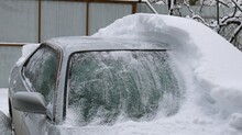 A Gray Car Covered In Snow And Half Cleared Of Snowdrift, The Consequences For Transport Of A Snow Storm And A Winter Disaster With A Blizzard And Snowfall, Cleaning A Vehicle From Fallen Snow