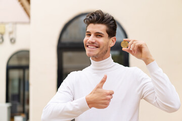 Wall Mural - Young caucasian man holding a Bitcoin at outdoors with thumbs up because something good has happened