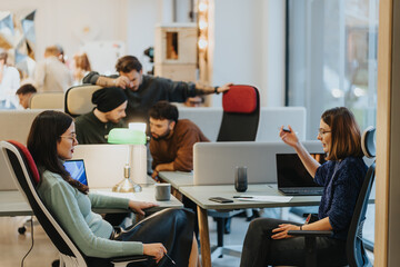 A focused view of two women conversing in a bustling co working environment, with other creative professionals collaborating in the background. The modern workspace fosters teamwork and shared ideas.