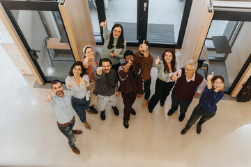 Wall Mural - Overhead view of a multicultural team smiling and showing thumbs up in an office setting, expressing teamwork and positivity at work.