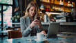 © MP Studio - woman is looking at her smartphone with a laptop open in front of her and a cup of coffee on the table, sitting in a cafe environment