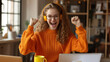 © Studio Nova - joyful young woman with curly hair, wearing glasses and an orange sweater, raising her fists in excitement while wearing headphones and sitting in front of a laptop