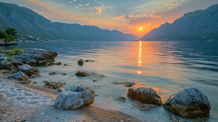 Poster -  Bay during sunset, the sky painted with hues of orange and pink, the sun setting behind the mountains, reflecting on the rippling waves and rocky shoreline 