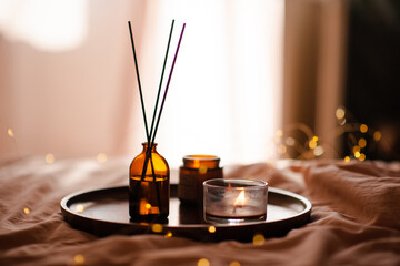 Wall Mural - Liquid home perfume in dark brown glass bottle and bamboo sticks with burning candles on wooden tray in bed room close up. Cozy hygge atmosphere.