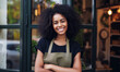 Portrait of young African-American curly woman small business owner of coffee shop standing at entrance wearing khaki apron and black shirt