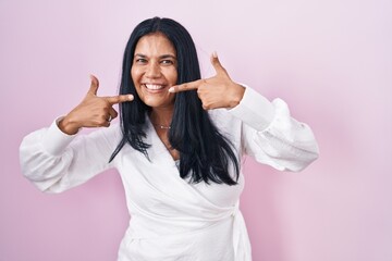 Poster - Mature hispanic woman standing over pink background smiling cheerful showing and pointing with fingers teeth and mouth. dental health concept.