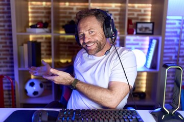 Poster - Middle age man with beard playing video games wearing headphones inviting to enter smiling natural with open hand