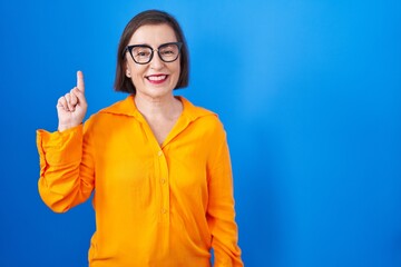 Wall Mural - Middle age hispanic woman wearing glasses standing over blue background showing and pointing up with finger number one while smiling confident and happy.