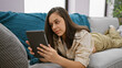 © Krakenimages.com - Relaxed young hispanic woman enjoys comfort of home, lying on sofa using touchpad, reflecting concentration of her beautiful face in restful web surf lifestyle, indoors