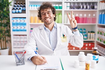 Canvas Print - Hispanic young man working at pharmacy drugstore smiling looking to the camera showing fingers doing victory sign. number two.