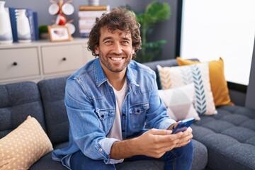 Wall Mural - Young hispanic man using smartphone sitting on sofa at home