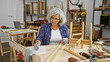 Mature woman measuring wood with tape in a cluttered carpentry workshop