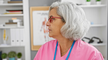 Mature woman healthcare professional with grey hair in pink scrubs inside medical clinic.