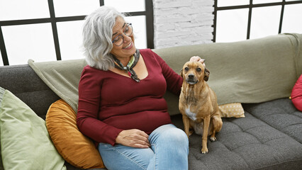 Canvas Print - Smiling mature woman with glasses pets her brown dog on a couch in a cozy living room.