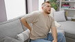 © Krakenimages.com - Young caucasian man with beard experiencing back pain at home, sitting on gray couch in a well-lit living room.