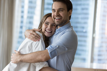 Close up portrait beautiful millennial spouses laughing, cuddling standing in modern apartment with city skyscrapers view. Homeowner family, bank loan, tenancy, start new life, cohabitation, marriage