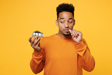 Wall Mural - Young man wear orange sweatshirt casual clothes hold in hand piece of cake dessert lick fingers isolated on plain yellow background studio. Proper nutrition healthy fast food unhealthy choice concept.