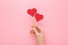 Young Adult Woman Hand Holding Two Red Paper Heart Shapes On Wooden Sticks On Light Pink Table Background. Pastel Color. Love Concept. Closeup. Top Down View.