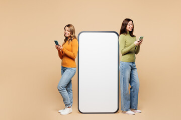 Wall Mural - Full body side view young friends two women they wear orange green shirt casual clothes together big huge blank screen mobile cell phone with area using smartphone isolated on plain beige background.