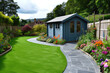 A general view of a back garden with artificial grass, grey paving slab patio, flower bed with plants, timber fences, blue shed, summer house garden timber outbuilding, with tropical garden