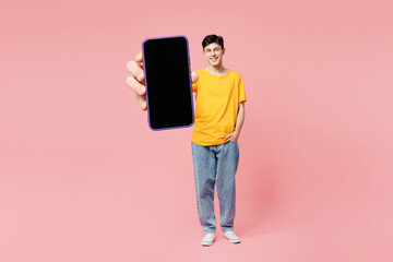 Wall Mural - Full body young smiling man he wears yellow t-shirt casual clothes show close up blank screen area mobile cell phone isolated on plain pastel light pink background studio portrait. Lifestyle concept.